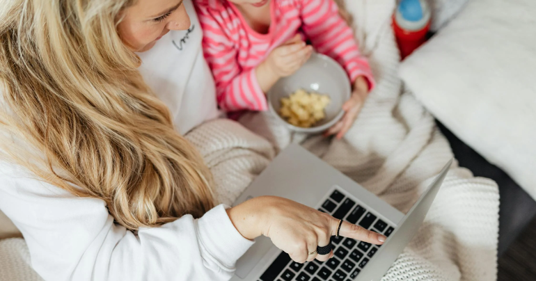Madre e hija disfrutan de un momento de entretenimiento frente a una laptop. La madre señala la pantalla mientras la niña observa con interés. Ambas comparten una experiencia agradable y divertida.