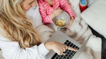Madre e hija disfrutan de un momento de entretenimiento frente a una laptop. La madre señala la pantalla mientras la niña observa con interés. Ambas comparten una experiencia agradable y divertida.
