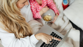 Madre e hija disfrutan de un momento de entretenimiento frente a una laptop. La madre señala la pantalla mientras la niña observa con interés. Ambas comparten una experiencia agradable y divertida.